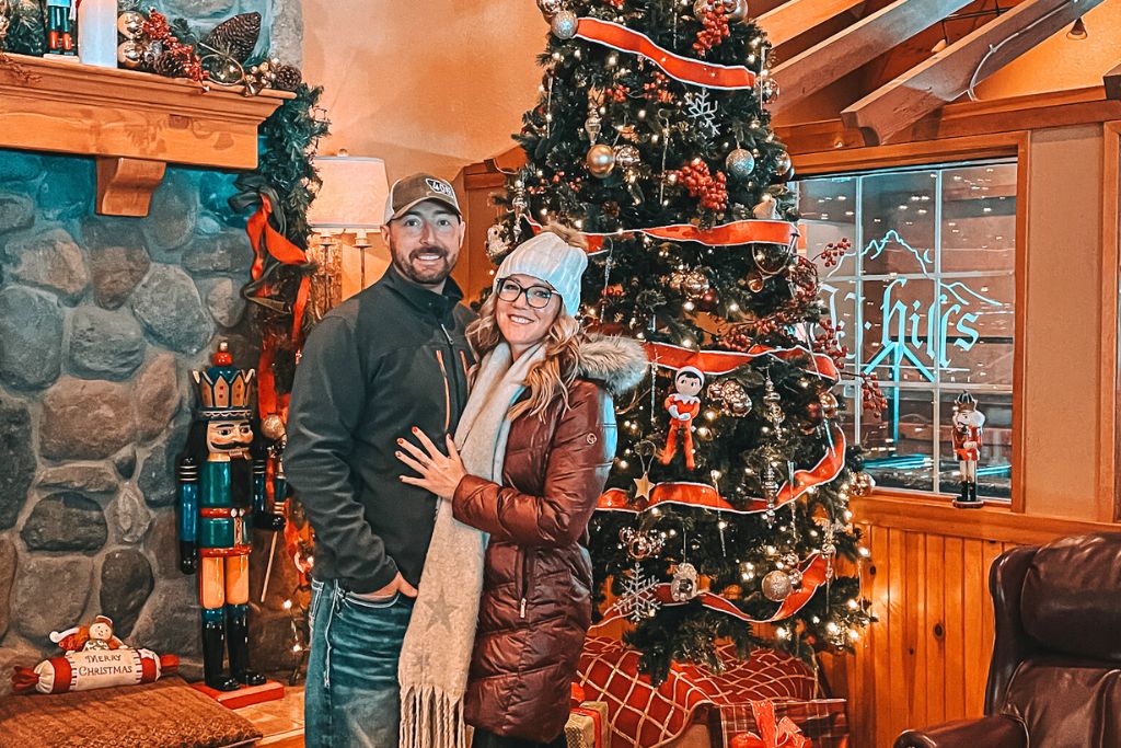 A cozy Christmas scene featuring Kate from Kate's Crossing Blog and her husband, standing in front of a beautifully decorated Christmas tree adorned with red ribbons, ornaments, and festive lights. The backdrop includes a stone fireplace with a Nutcracker and holiday decor, adding to the warm and festive atmosphere. The setting, with its rustic charm and holiday cheer, creates a welcoming environment perfect for celebrating Christmas in a cozy, lodge-like space.