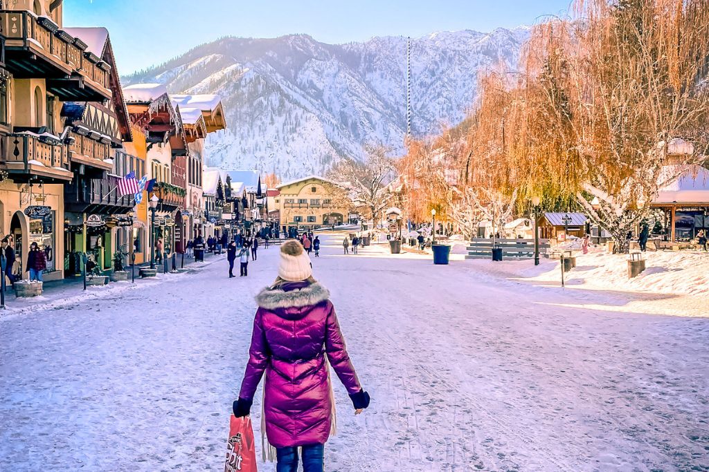 A winter scene in Leavenworth, with Kate from Kate's Crossing Blog walking down a snowy street lined with Bavarian-style buildings. The surrounding mountains, covered in snow, create a breathtaking backdrop. Kate, dressed warmly in a red coat and hat, is carrying a shopping bag, adding to the festive, holiday shopping atmosphere. The scene is bright and cheerful, with other people enjoying the picturesque setting, perfect for a winter getaway in one of the best places to stay in Leavenworth for Christmas.