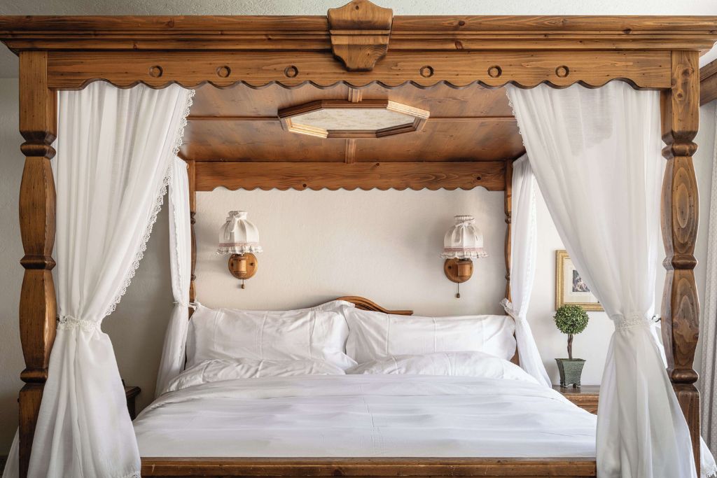 A cozy room at the Enzian Inn, featuring a rustic wooden canopy bed with intricate detailing. The bed is draped with light white curtains and dressed in crisp white linens, creating a charming, traditional feel. Two vintage-style lamps hang on the wall above the bed, enhancing the room's warm and inviting atmosphere. A small potted plant adds a fresh touch to the wooden side table next to the bed.