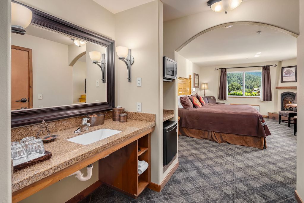 A spacious room at the Bavarian Lodge in Leavenworth, featuring a large bed with dark brown linens and vibrant accent pillows. The room is divided by an archway, leading from the vanity area with granite countertops and a large mirror into the bedroom space. A window offers a scenic view of lush green mountains, while a small fireplace adds a cozy touch near the seating area. The room also includes a microwave and mini-fridge, blending convenience with comfort.