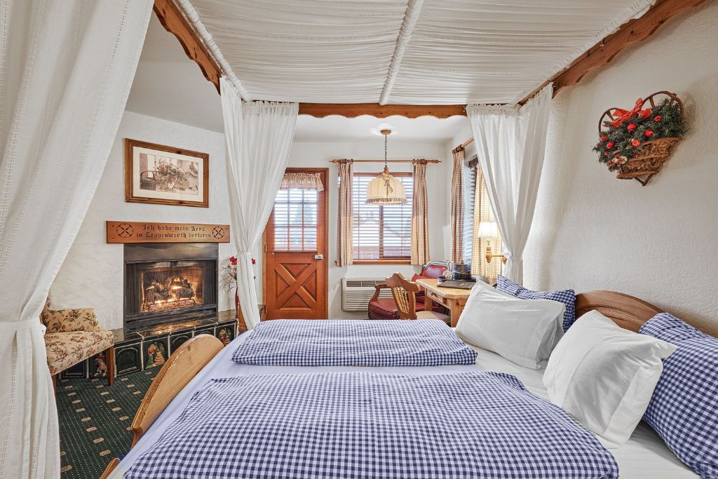 A charming room at Hotel Pension Anna, featuring a wooden canopy bed with blue and white checkered bedding and draped white curtains. The room has a warm, festive feel, with a cozy fireplace framed by traditional decor and a floral-patterned bench nearby. A small desk and chair are positioned by the window, allowing natural light to flow in, and a holiday wreath hangs on the wall. The room is accented with rustic wood details, including a door that opens to a scenic view.