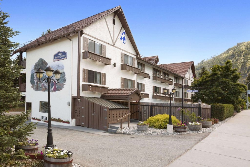 The exterior of Howard Johnson by Wyndham in Leavenworth, designed with a traditional Alpine style, featuring brown wooden shutters and a tiled roof. A mural on the side of the building adds a charming, Bavarian touch. Flower pots line the walkway, adding vibrant color to the scene, and a vintage-style lamppost enhances the quaint, picturesque feel. The hotel is nestled against a backdrop of green mountains, creating a peaceful, scenic atmosphere.