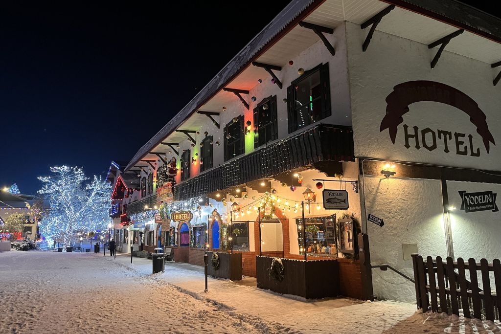 A festive, snow-covered street in Leavenworth, highlighting one of the best places to stay in Leavenworth for Christmas. The building, styled with Bavarian architecture, is adorned with colorful Christmas lights and holiday decorations, creating a warm and inviting holiday ambiance. Twinkling lights illuminate the surrounding trees, while the snow enhances the magical, wintery atmosphere, perfect for a cozy Christmas getaway.