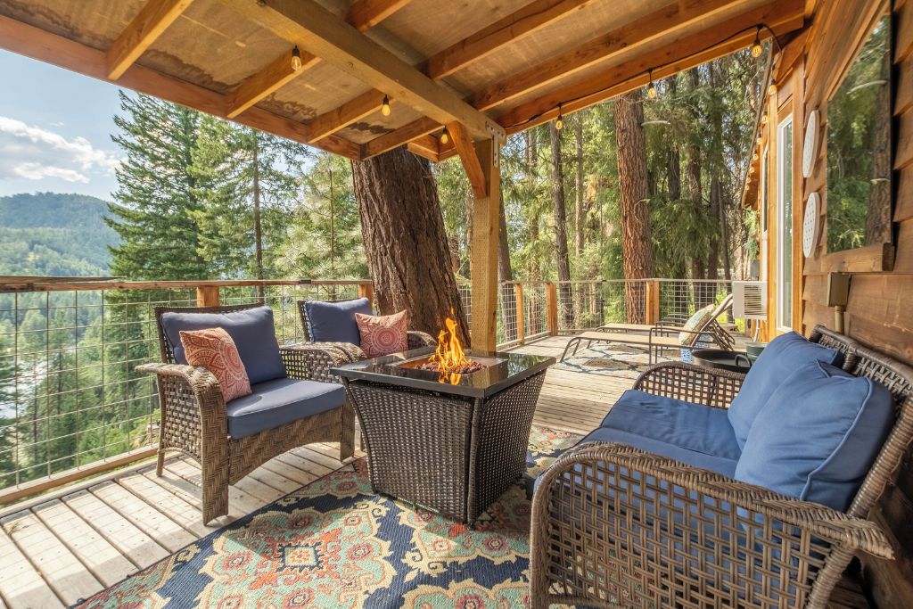 The outdoor deck of the Das Tree Haus vacation rental near Leavenworth. The deck is furnished with wicker chairs featuring blue cushions and decorative throw pillows, arranged around a gas firepit for warmth and ambiance. The area is surrounded by tall trees, providing a serene, forested view, with a distant glimpse of mountains in the background. The deck has a rustic, cabin-like charm, enhanced by the wooden beams overhead and a cozy, patterned outdoor rug.