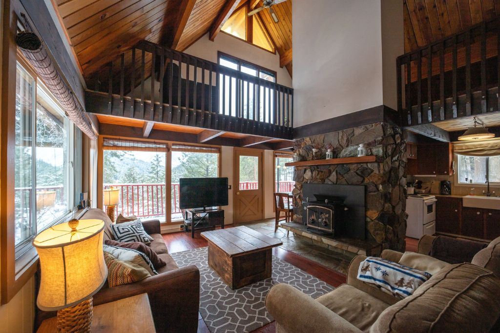 Cozy interior of a Leavenworth cabin, featuring a warm, rustic living area with high, vaulted ceilings and exposed wooden beams. A large stone fireplace and wood-burning stove serve as the focal point, adding to the cabin's inviting atmosphere. The space includes comfortable seating, a wooden coffee table, and a TV, with large windows and a sliding door that offer views of the surrounding forest. A loft area overlooks the living room, and the adjacent kitchen is equipped with modern appliances, blending comfort with a rustic charm.