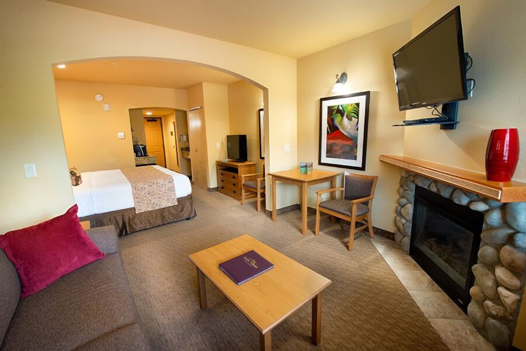 A spacious room at Icicle Village Resort, featuring a cozy seating area with a couch and a table, a bed with a light-colored throw, and a fireplace framed by stone. The room includes a flat-screen TV mounted above the fireplace, a small desk and chair, and warm, neutral tones that create a welcoming ambiance. A bright red vase and colorful wall art add touches of color to the space.