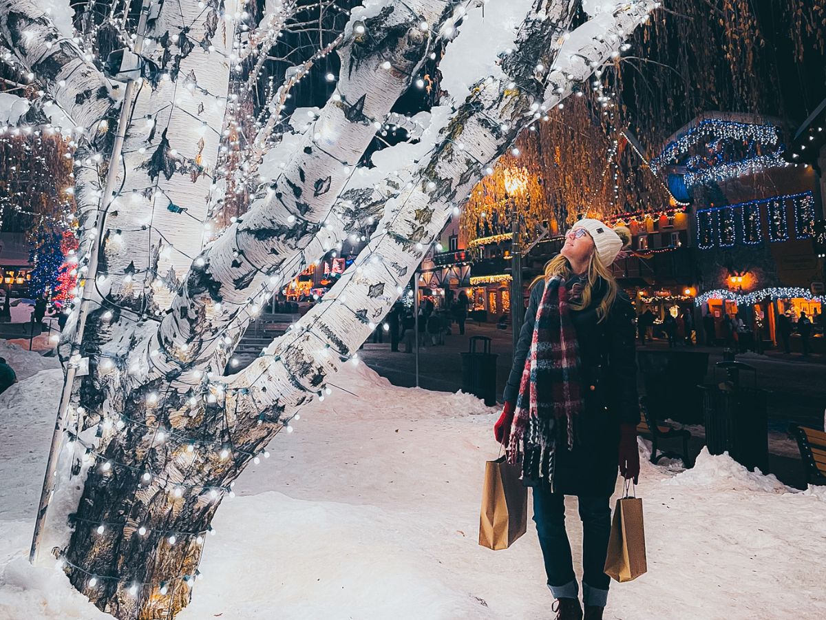 Kate from the Kate's Crossing blog, standing beside a snow-covered tree adorned with sparkling holiday lights in Leavenworth, Washington. She is bundled up in a winter coat, scarf, and beanie, holding shopping bags, while gazing up at the festive lights, capturing the essence of the town’s enchanting holiday season. The scene illustrates the joy of visiting Leavenworth in winter, with its beautiful decorations and magical ambiance, making it a perfect time to explore.