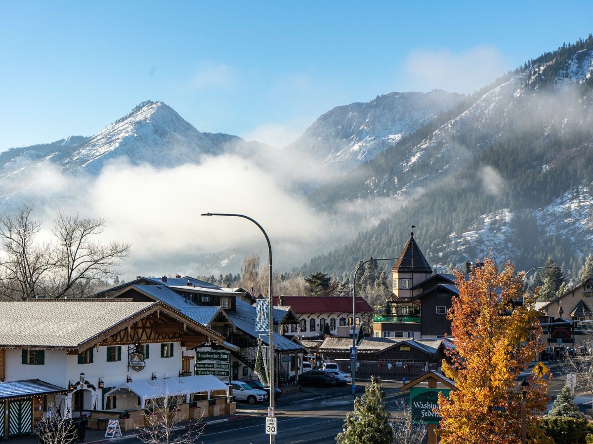 a stunning view of Leavenworth, Washington, on a crisp day with mist rolling over the snow-dusted mountains in the background. The Bavarian-style buildings in the foreground are accented by autumn foliage, with a golden tree standing out against the cool mountain scenery. This serene blend of fall and early winter landscapes highlights why this transitional period might be considered the best time to visit Leavenworth, combining the beauty of changing seasons.