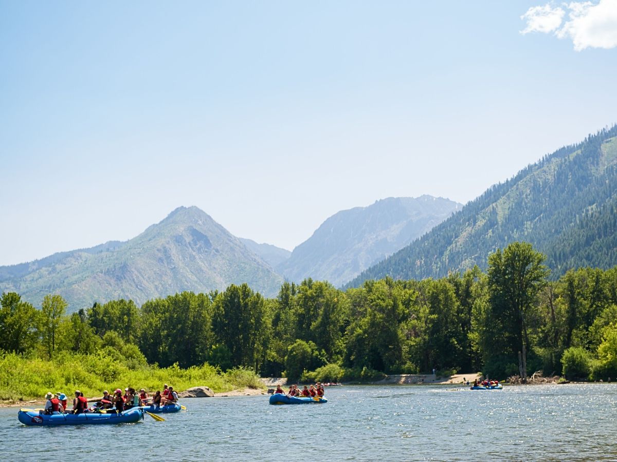 a summer day in Leavenworth, Washington, with groups of people rafting on a calm river surrounded by lush greenery. The backdrop features the towering Cascade Mountains under a clear, blue sky, creating a picturesque outdoor adventure scene. Summer in Leavenworth is ideal for water activities and enjoying nature, which makes it one of the best times to visit for those seeking outdoor recreation and scenic beauty.