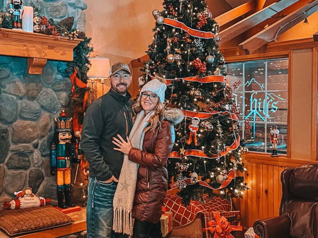 Kate from Kate's Crossing Blog and her husband standing in front of a beautifully decorated Christmas tree inside the cozy Icicle Village Resort in Leavenworth, Washington. The room has a warm and festive ambiance, with a stone fireplace adorned with garlands and a nutcracker figurine beside it. They are dressed in winter clothing, smiles warmly, capturing the holiday spirit in this charming winter destination. 