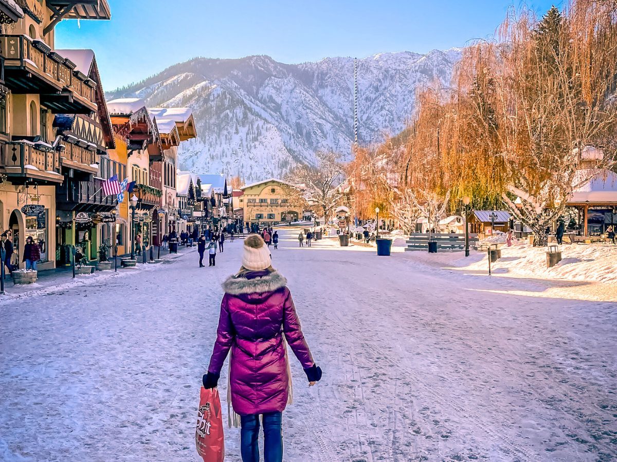 Kate from the Kate's Crossing blog walking down a snow-covered street in Leavenworth, Washington. She is wearing a purple winter coat with a fur-lined hood and holding a shopping bag, while the town’s charming Bavarian-style buildings and snow-capped mountains provide a picturesque backdrop. The clear sky and the soft glow of sunlight on the winter landscape make this a serene and beautiful moment.