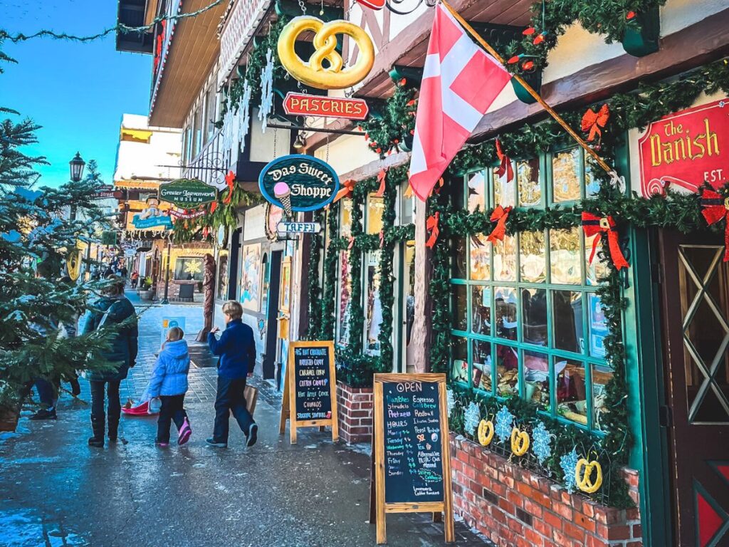 A festive street in Leavenworth, Washington, with shops decorated for the holiday season. The storefronts are adorned with garlands, red bows, and Christmas lights, and signs for pastries and local treats hang overhead. A family walks along the sidewalk, dressed warmly for winter, adding to the cozy and inviting atmosphere of the town. This scene captures the charm of Leavenworth during the holiday season.