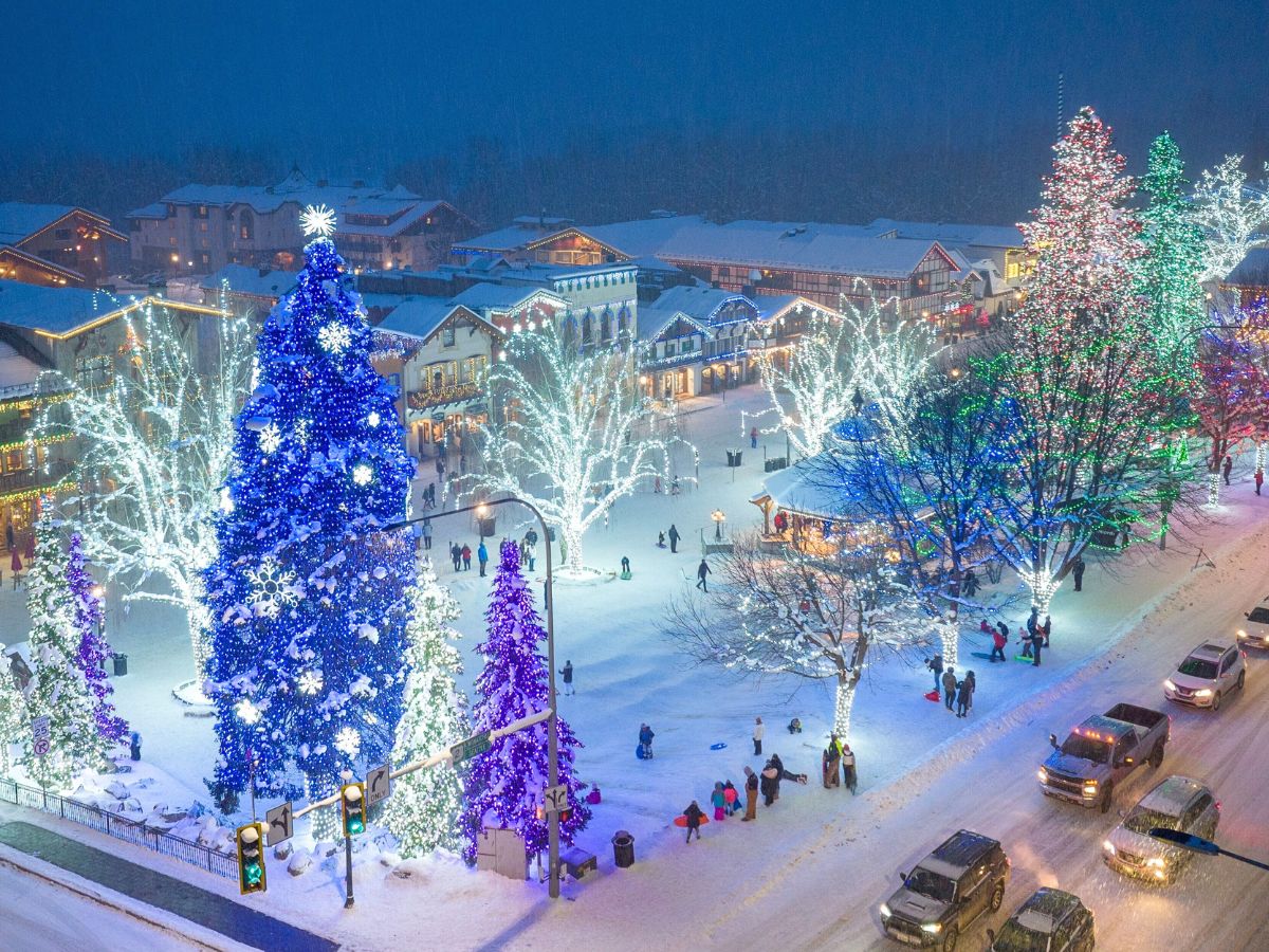 A breathtaking winter scene in Leavenworth, Washington, during the holiday season. The town square is illuminated with vibrant Christmas lights, featuring a large blue Christmas tree adorned with snowflakes and other festive decorations. Surrounding trees are lit up with twinkling lights, while families and visitors enjoy the snow-covered streets below. The scene showcases Leavenworth's magical winter wonderland.