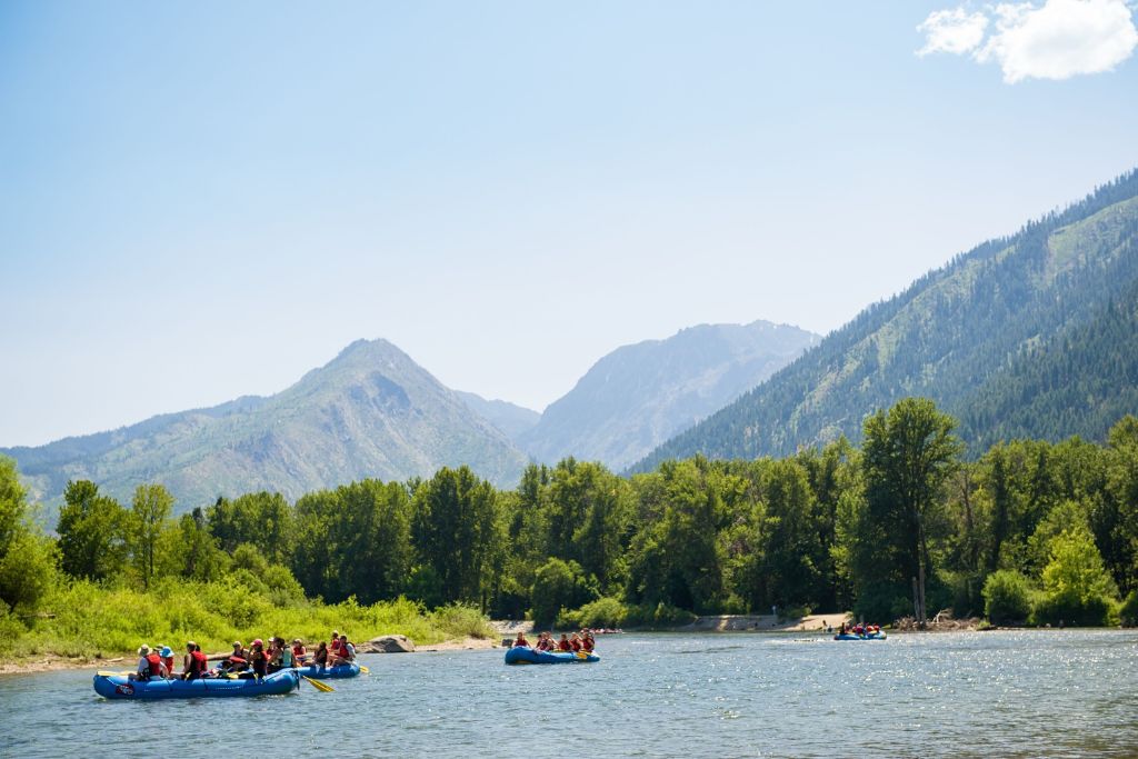 Groups of people enjoy white water rafting on a calm section of a river near Leavenworth, Washington. They paddle in blue rafts while surrounded by lush green trees, with towering mountains in the background under a clear blue sky, creating a scenic outdoor adventure setting.