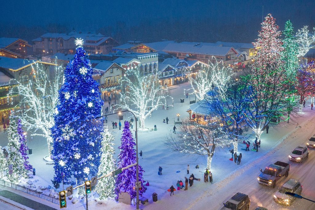 A breathtaking nighttime view of downtown Leavenworth, Washington, during the holiday season. The town square is illuminated with vibrant Christmas lights, including a towering blue Christmas tree decorated with snowflake ornaments. Colorfully lit trees and a snow-covered gazebo add to the festive ambiance, while people walk through the snowy streets and cars drive past the beautifully decorated town.