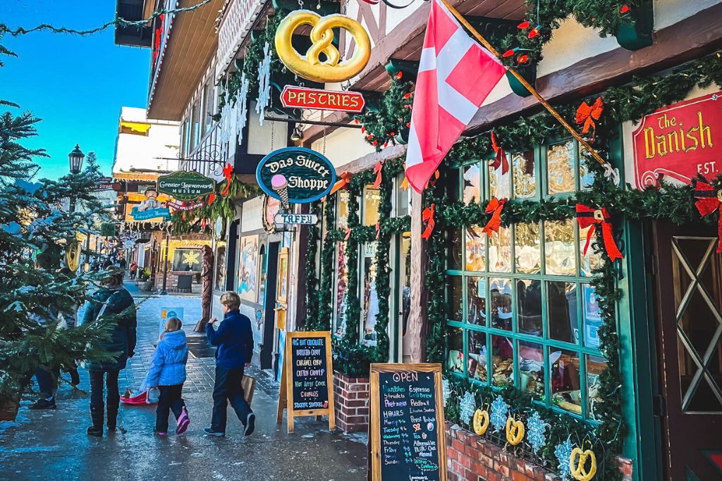 A festive street in Leavenworth, Washington, decorated for the holidays with garlands, wreaths, and red bows. Shop signs, including 'Das Sweet Shoppe' and 'The Danish,' hang along the storefronts, while a Danish flag flutters overhead. People, including children bundled in winter coats, stroll past, enjoying the charming atmosphere and window displays of the pastry and candy shops.