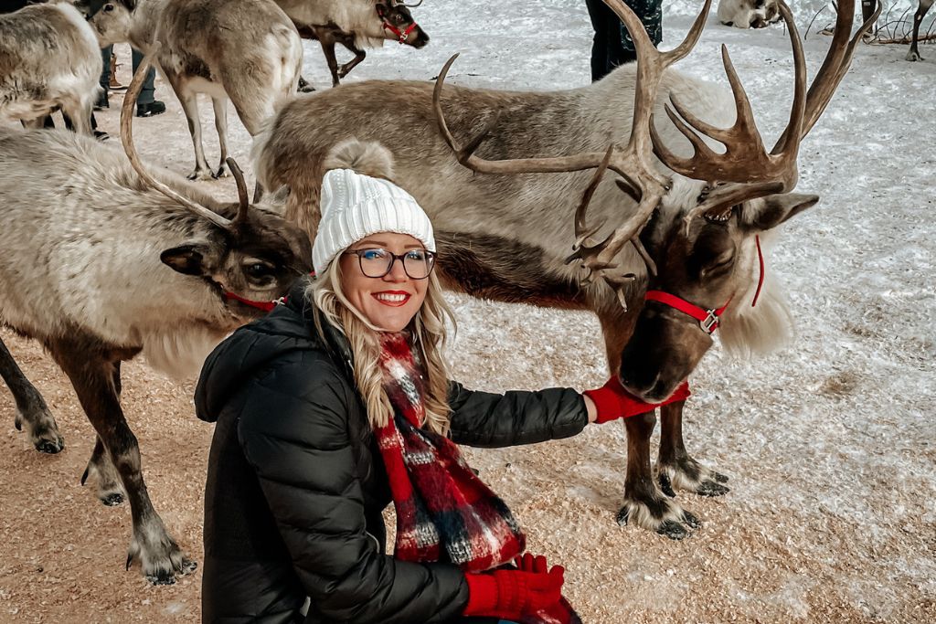 Kate from Kate’s Crossing Blog kneels beside a reindeer, petting it gently, while surrounded by several other reindeer. She is bundled up in a white beanie, red scarf, and gloves, smiling in the snowy outdoor setting. The reindeer wear red harnesses, adding to the winter wonderland vibe of the scene.