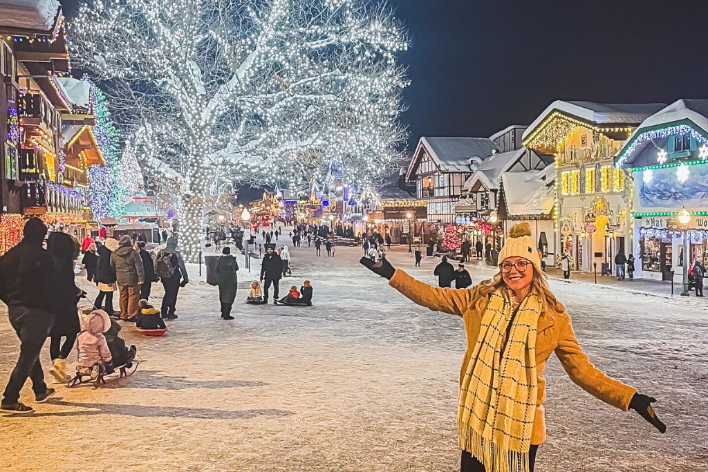 Kate from Kate’s Crossing Blog poses in Leavenworth, Washington, at night during the winter season. She is smiling, dressed in a yellow coat, scarf, and beanie, with the festive, brightly lit street and trees twinkling behind her. People are seen enjoying the holiday atmosphere, with some pulling sleds, as the alpine-style buildings glow with colorful lights.