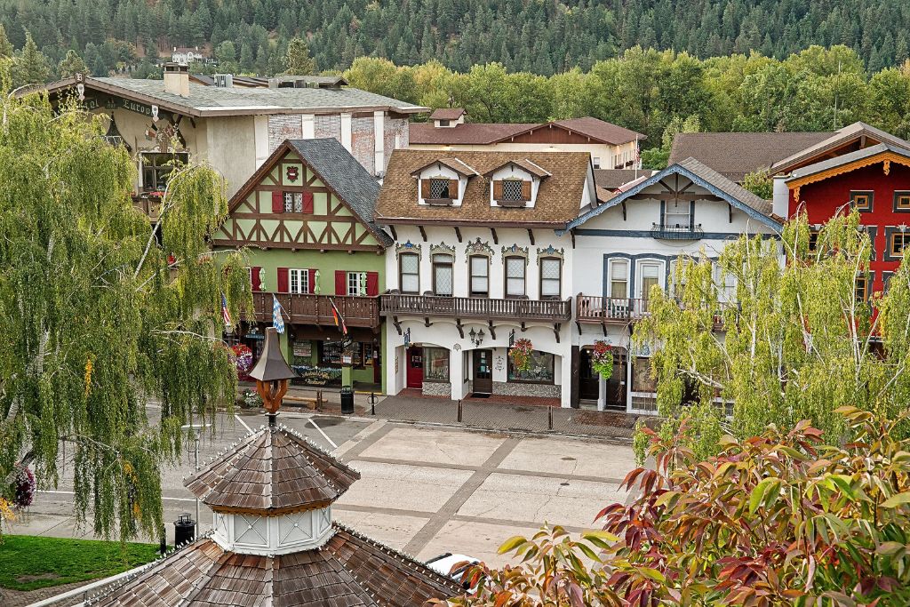 An overhead view of Leavenworth, Washington, showcasing its distinct alpine-style buildings with timber-framed facades in green, white, and red. The scene is framed by lush green trees in the foreground and background, with a gazebo rooftop visible in the lower part of the image. The charming architecture contrasts with the surrounding natural landscape, adding to the town's Bavarian-inspired appeal.