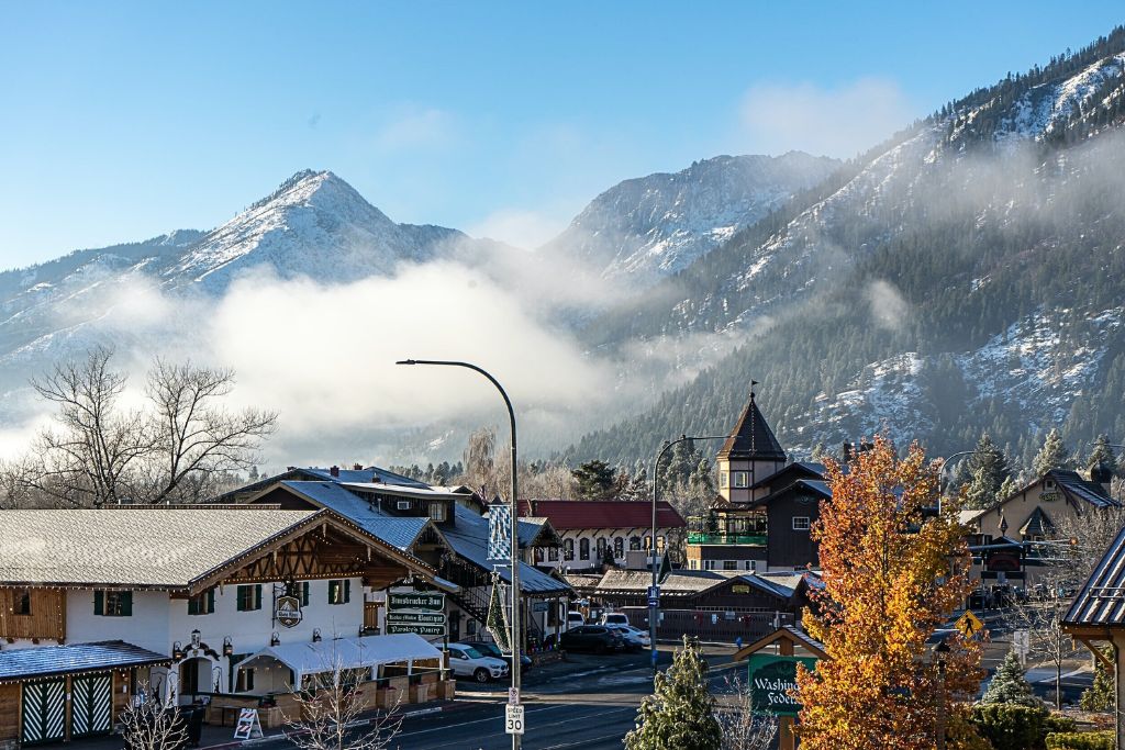 A scenic view of Leavenworth, Washington, with snow-dusted alpine-style buildings in the foreground and towering, snow-covered mountains in the background. Low clouds and mist drift around the mountains, creating a serene and picturesque atmosphere. The fall foliage adds a splash of color to the scene, contrasting with the snowy peaks and clear blue sky.