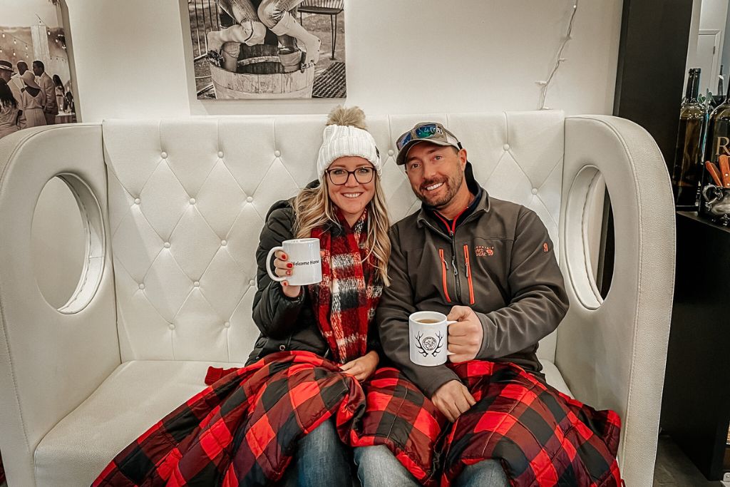 Kate from Kate’s Crossing Blog and a companion sit together on a plush, white, tufted bench, both wrapped in a red and black plaid blanket. They are smiling and holding mugs, with Kate's mug displaying the words 'Welcome Home.' The cozy indoor setting, along with their winter attire, creates a warm and inviting atmosphere.