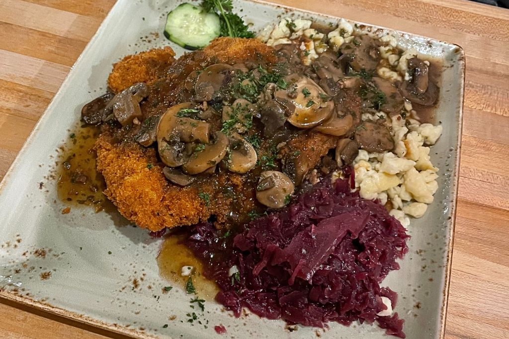 A plate of German-inspired cuisine featuring a crispy schnitzel topped with mushroom gravy, accompanied by red cabbage, spaetzle, and a sliced cucumber garnish. The dish is served on a large square plate, showcasing a hearty and flavorful meal.