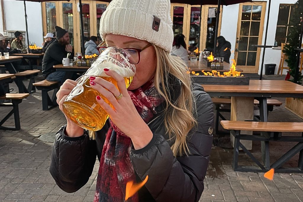 Kate from Kate’s Crossing Blog drinks from a large glass stein of beer while seated at an outdoor beer garden in Leavenworth, Washington. She’s bundled up in a winter hat, scarf, and jacket, with a cozy firepit visible in the background among wooden picnic tables, creating a warm, inviting atmosphere for enjoying the cold weather.