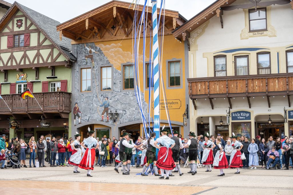 A traditional Bavarian maypole dance taking place in the town square of Leavenworth, Washington. Dancers in traditional German attire, with men in lederhosen and women in dirndls, perform around a tall pole with blue and white ribbons. A crowd of onlookers, standing in front of colorful, alpine-style buildings, enjoys the lively cultural performance.