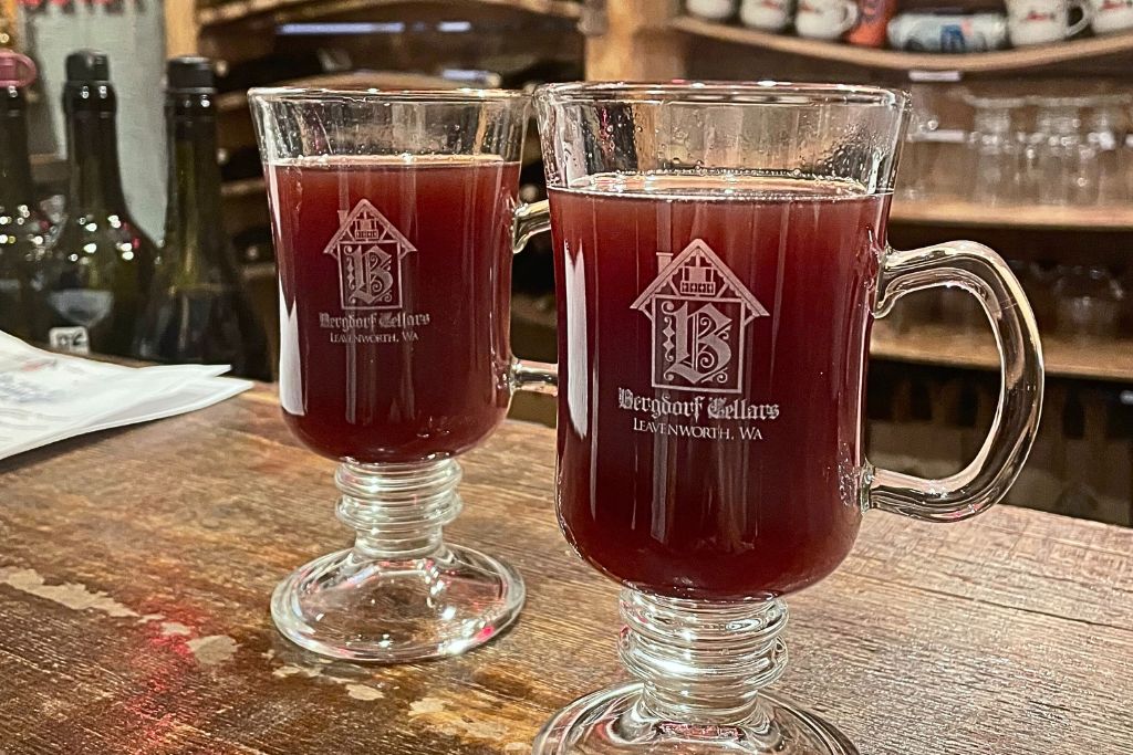 Two glass mugs of mulled wine served at Bergdorf Cellars in Leavenworth, Washington. The rich, red beverage is set on a rustic wooden counter, with the mugs featuring the Bergdorf Cellars logo etched on the front. The cozy, inviting atmosphere of the wine bar is hinted at in the background.