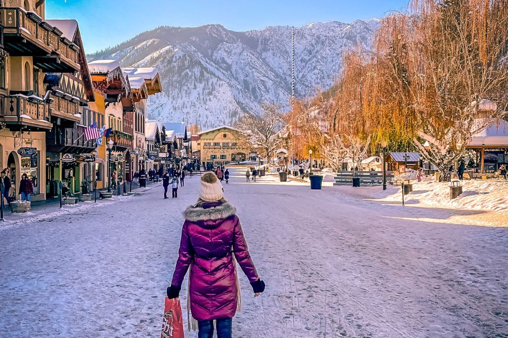 Kate from Kate’s Crossing Blog walks down a snow-covered street in Leavenworth, Washington. She’s wearing a red winter coat and beanie, while picturesque, alpine-style buildings line both sides of the street. The scene is set against a backdrop of snow-capped mountains under a clear blue sky, capturing the charm of the wintery town.