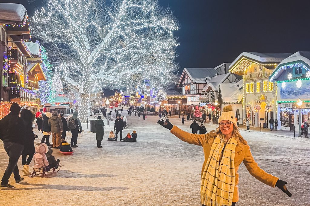 A lively winter scene in Leavenworth, Washington, during February. The town is lit up with festive lights, with snow-covered streets and buildings adding to the magical atmosphere. Kate from Kate's Crossing Blog, enjoying the winter festivities, poses in the foreground, smiling in her warm coat and scarf, while families and visitors stroll and sled behind her. February in Leavenworth, with its snowy charm and bright lights, is a wonderful time to visit for a cozy and picturesque winter experience.