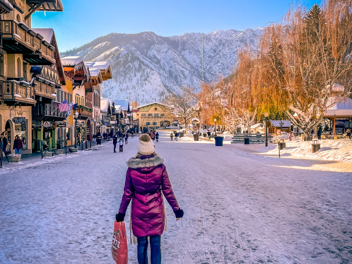 The image shows a scenic winter day in downtown Leavenworth, Washington. Kate, dressed warmly in a maroon coat and white knit hat, walks down a snow-covered street lined with Bavarian-style buildings. They are carrying a red shopping bag, and the scene is framed by snowy mountains in the background under a clear blue sky. The street is bustling with people, and the charming town is illuminated by the soft golden glow of the afternoon sun, highlighting the festive, alpine atmosphere of Leavenworth.