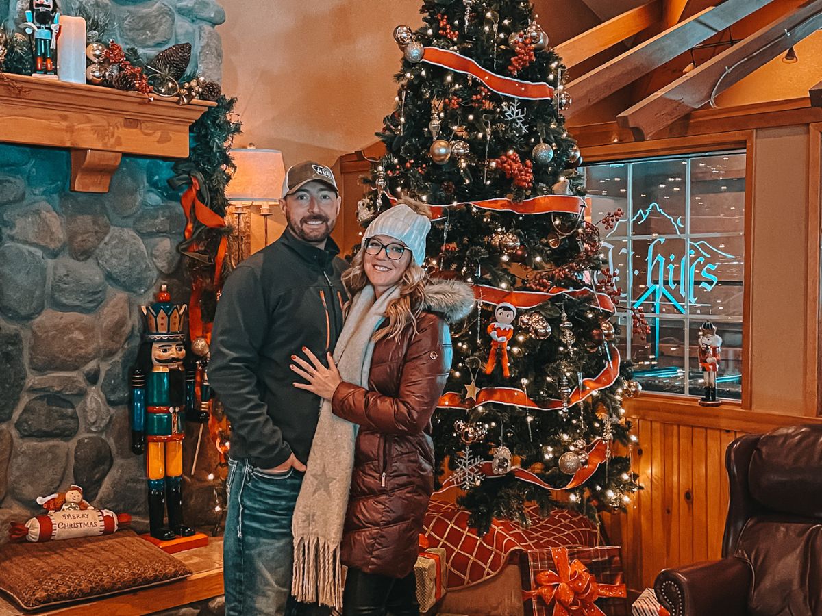 Kate from Kate's Crossing blog and her companion posing in front of a beautifully decorated Christmas tree inside a cozy, festive setting. The tree is adorned with red ribbon, ornaments, and lights, with wrapped presents placed beneath it. To the left, a stone fireplace is decorated with holiday garlands and a nutcracker figure, adding to the Christmas ambiance. The couple is warmly dressed in winter clothing, smiling as they stand together, embracing the holiday spirit. A sign reading "Merry Christmas" sits on the hearth, further enhancing the festive mood.