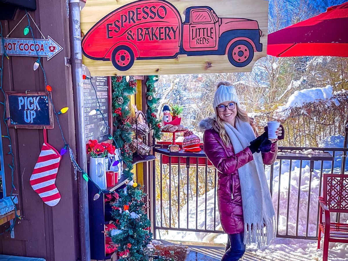 Kate from Kate's Crossing blog standing outside an espresso and bakery stand in a festive, winter setting. She is smiling and holding a hot beverage, dressed warmly in a pink coat, scarf, gloves, and a knit beanie. The stand is decorated with holiday-themed items like a candy cane-striped stocking, Christmas lights, and a wooden sign for "Little Reds Espresso & Bakery," with snowy scenery in the background. A small chalkboard sign reading "Pick Up" and another advertising hot cocoa add to the cozy, inviting atmosphere.