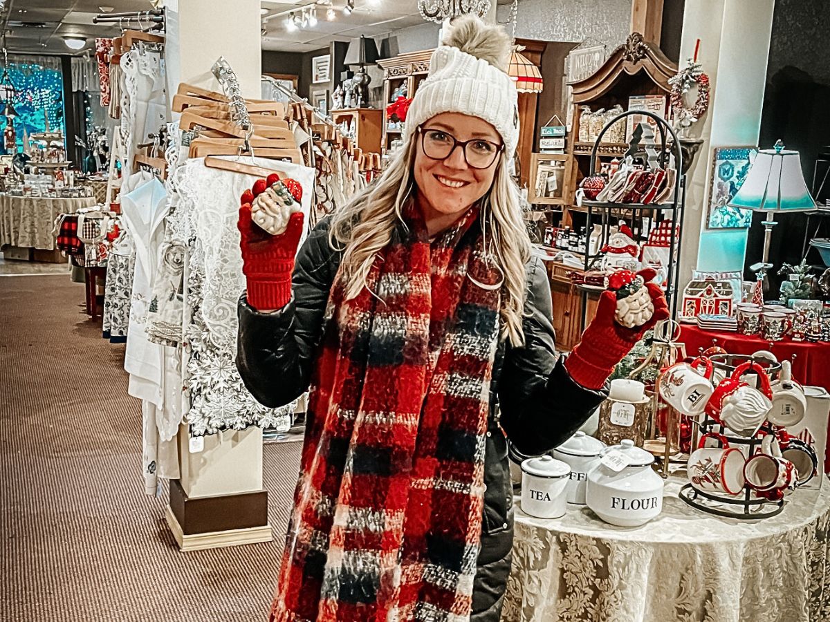 Kate from Kate's Crossing blog inside a cozy gift shop in Leavenworth, Washington. She is bundled up in a winter outfit, including a white knit beanie, red gloves, and a plaid scarf. Kate is smiling and holding up two festive holiday ornaments in her hands, while the shop around her is filled with Christmas decor, ornaments, and other seasonal goods. The warm lighting and holiday decorations add to the festive shopping atmosphere.