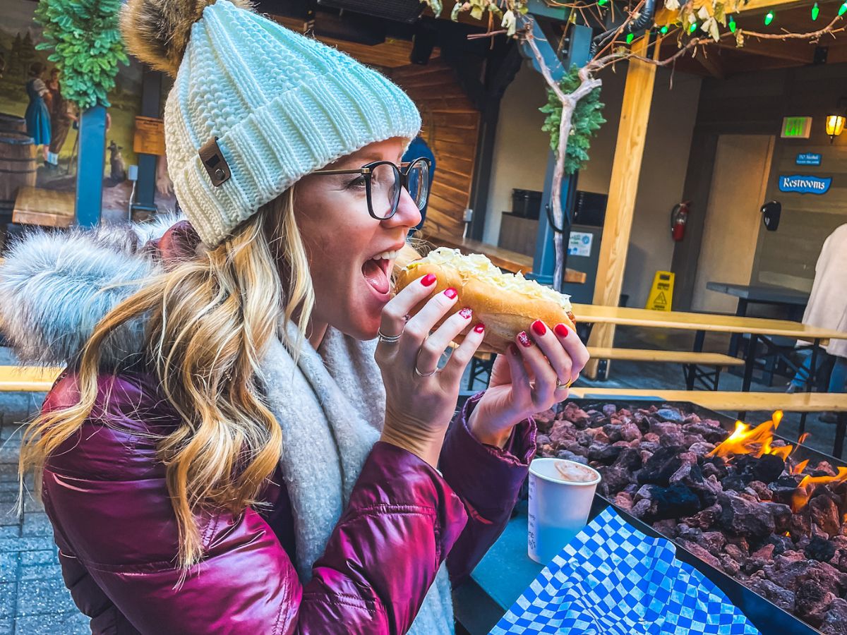 Kate from Kate's Crossing blog enjoying a meal outdoors in Leavenworth, Washington. She is dressed warmly in a white knit hat, a scarf, and a maroon winter jacket with a fur-lined hood. Kate is about to take a bite of a sausage in a bun topped with sauerkraut, while sitting by a cozy firepit. The scene has a festive, rustic atmosphere, with outdoor seating and holiday decor visible in the background. Her bright red nails and winter attire add to the cozy, wintertime vibe.