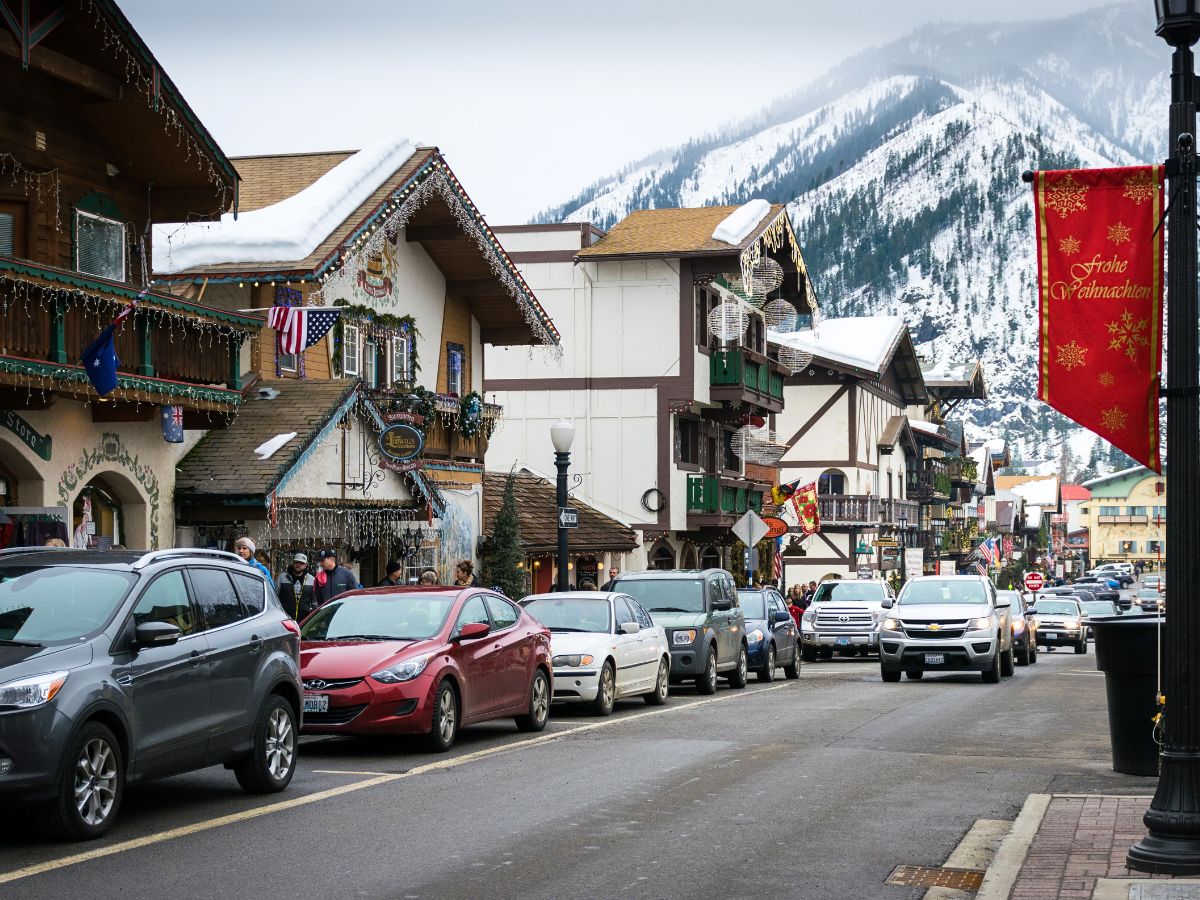 A lively street scene in downtown Leavenworth, featuring Bavarian-style buildings adorned with festive holiday lights and decorations. Cars line the street, and pedestrians stroll past the charming shops, with snow-covered mountains towering in the background. A red banner with the German phrase "Frohe Weihnachten" (Merry Christmas) adds a festive touch to the wintery atmosphere.