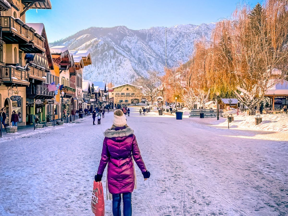  Kate from Kate's Crossing Blog in a marron winter coat walks down a snow-covered street in downtown Leavenworth, surrounded by Bavarian-style buildings. The scene is bright and vibrant, with snow-dusted trees and mountains in the background under a clear blue sky. People stroll and shop along the festive street, enjoying the picturesque winter atmosphere.