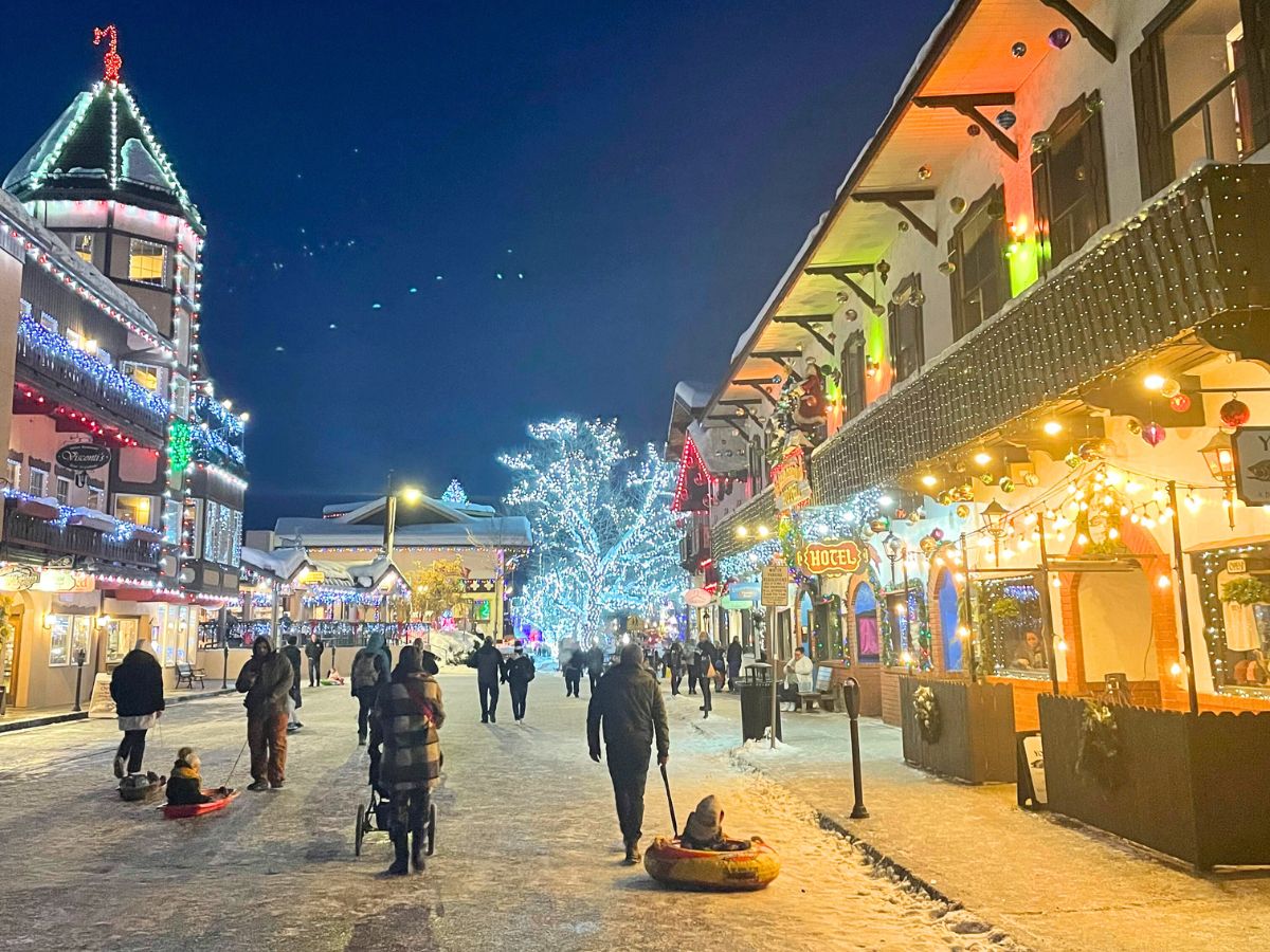 A festive winter night in downtown Leavenworth, with Bavarian-style buildings adorned in colorful holiday lights. People stroll along the snow-covered street, some pulling children on sleds, while others enjoy the bright, cheerful atmosphere. The scene is illuminated by sparkling lights on buildings and a large tree, with the dark night sky providing a beautiful contrast to the vibrant decorations.