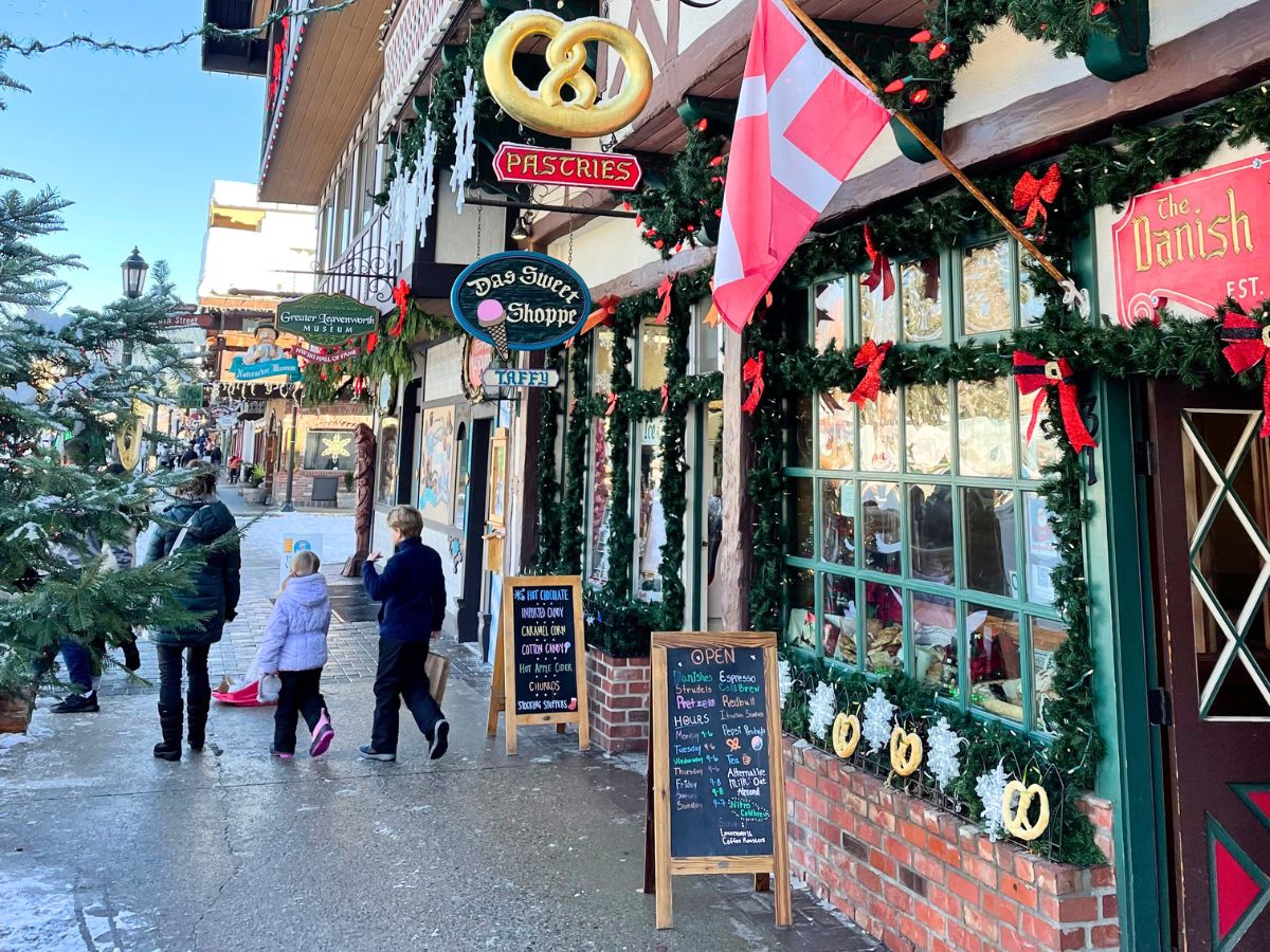 A charming street in downtown Leavenworth, featuring festive holiday decorations on shopfronts. Signs for "Das Sweet Shoppe" and "The Danish" bakery hang above the entrance, while garlands and red bows adorn the windows. A few people, including a child in a purple coat, walk down the sidewalk, enjoying the Bavarian-themed shops. A large pretzel sign adds to the cozy and inviting atmosphere of this wintery scene.