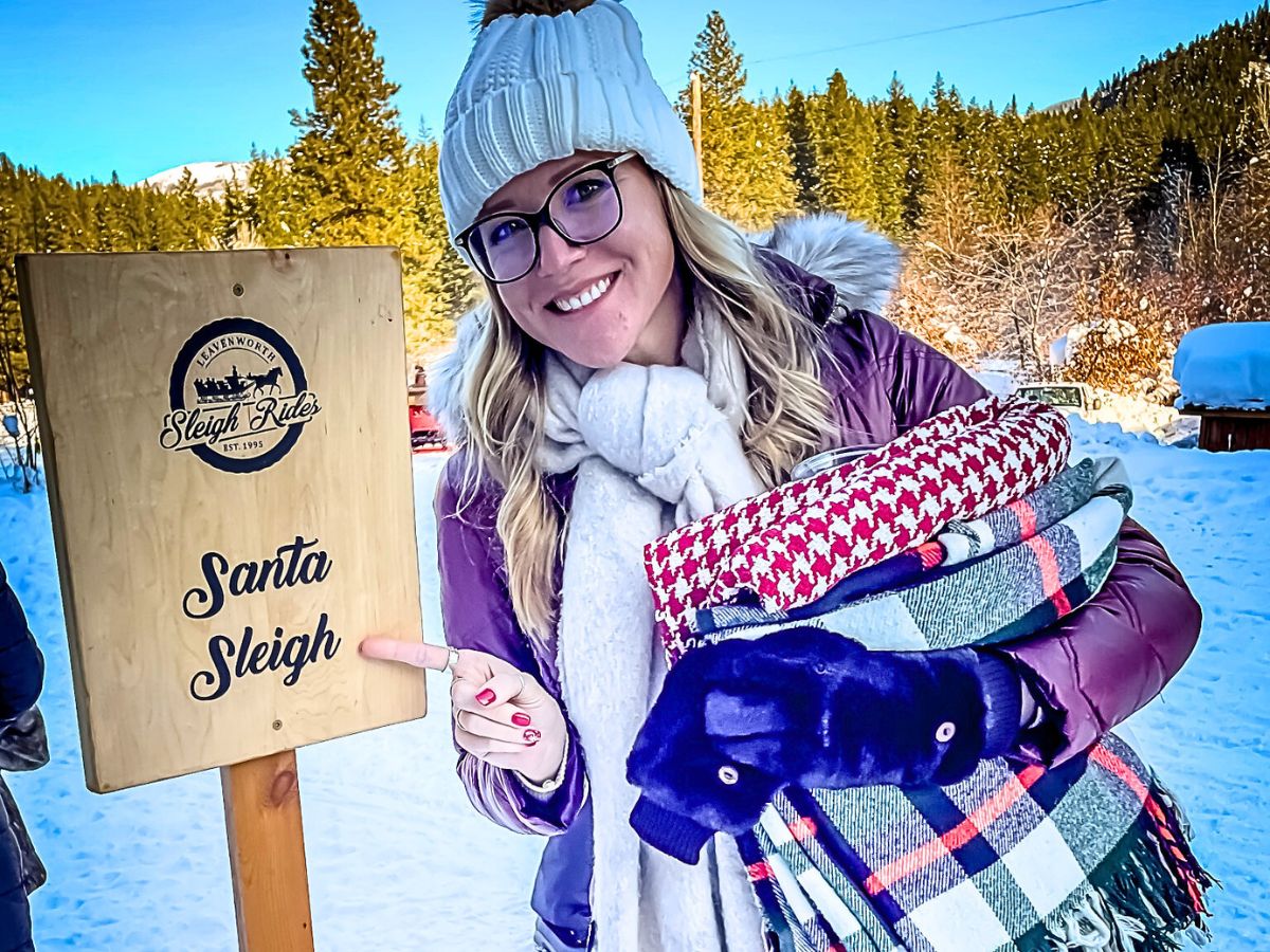 Kate from Kate's Crossing Blog dressed in winter attire holds several cozy blankets while standing next to a wooden sign that reads "Santa Sleigh" with a logo for Leavenworth Sleigh Rides. The snowy background features pine trees and clear blue skies, creating a festive and inviting winter atmosphere. The woman points to the sign, excited for the sleigh ride experience.