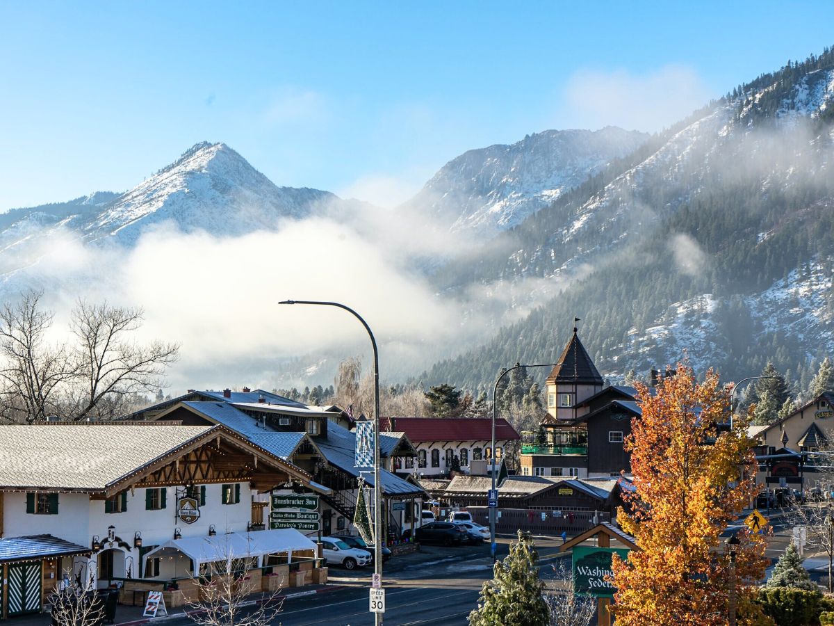 A picturesque view of downtown Leavenworth with Bavarian-style buildings set against the backdrop of snow-capped mountains and low-lying clouds. The town features classic Alpine architecture, with signs and colorful fall foliage adding to the charm. Mist hovers over the surrounding peaks, creating a crisp, tranquil atmosphere under a bright blue sky.