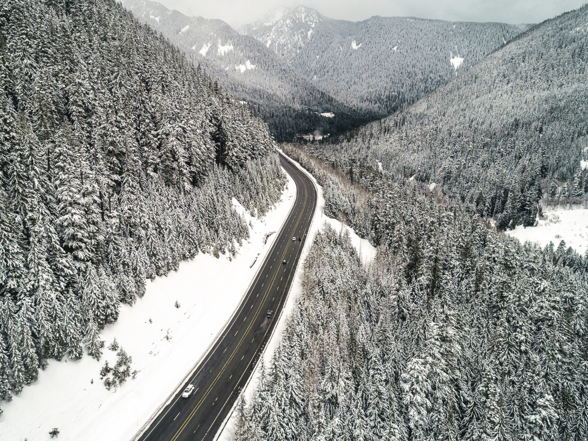 Aerial view of the snow-covered Stevens Pass along the Seattle to Leavenworth route, showcasing a highway cutting through dense, frosted evergreen forests in the Cascade Mountains. A few cars navigate the road as the surrounding landscape is blanketed in snow, creating a serene winter scene with towering mountains in the distance.