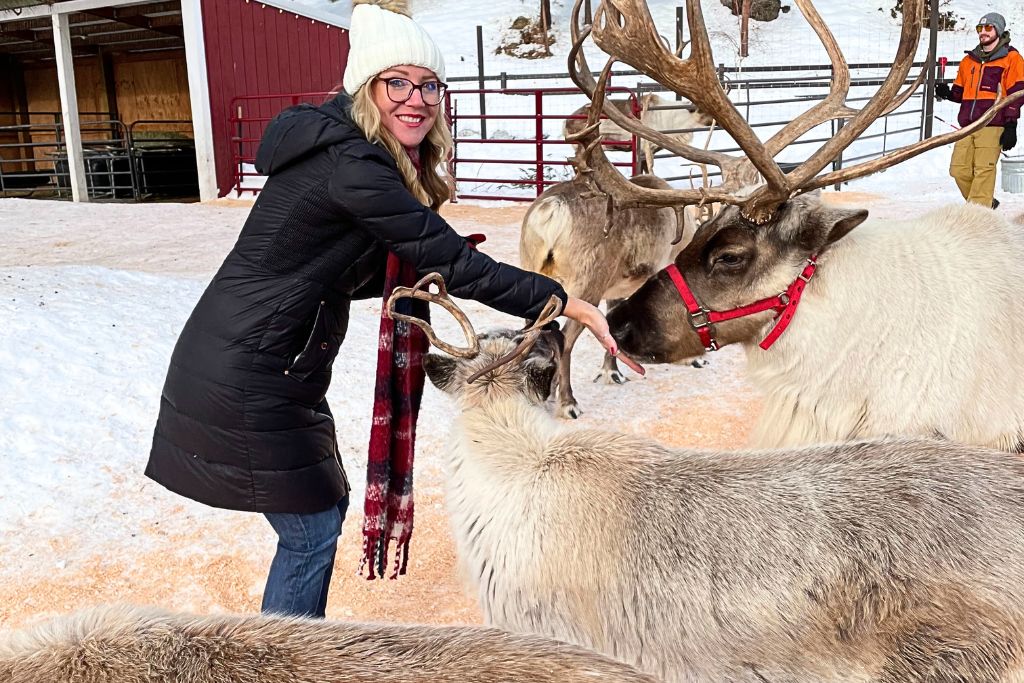 Kate from Kate's Crossing Blog is seen petting a group of reindeer, including one with a red halter, at a snowy reindeer farm. Dressed in a long black winter coat, white knit hat, and plaid scarf, she smiles while interacting with the animals.