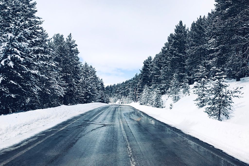 A serene winter road surrounded by snow-covered pine trees, with the road itself slightly wet from melted snow. The peaceful, scenic drive through the snow-covered forest offers a tranquil winter experience.