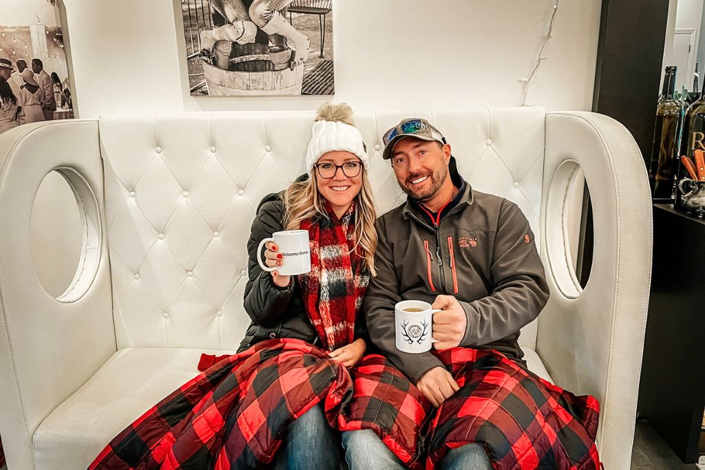 Kate from Kate's Crossing Blog and a companion sitting on a cozy white sofa, both holding mugs and wrapped in a red plaid blanket. They are enjoying warm drinks in a comfortable indoor setting, smiling at the camera.