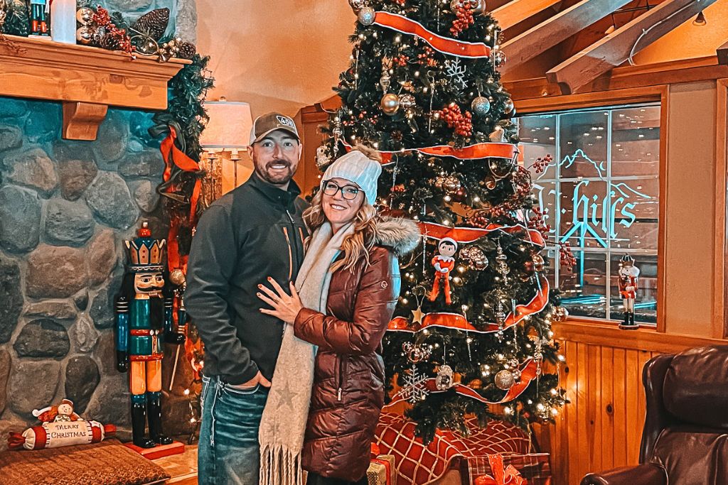 Kate from Kate's Crossing Blog and a companion posing in front of a beautifully decorated Christmas tree inside a cozy, festive space. The room is adorned with holiday decorations, including a nutcracker and garland above a stone fireplace.