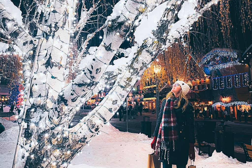 Kate from Kate's Crossing Blog stands beneath a snow-covered tree adorned with sparkling white lights, gazing up at the festive display. The backdrop features brightly lit buildings, creating a magical winter atmosphere in downtown Leavenworth.