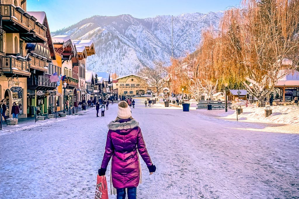 A winter scene in downtown Leavenworth with Kate from Kate's Crossing Blog in a purple coat walking down a snow-covered street, holding a shopping bag. The picturesque Bavarian-style buildings line the street, and snow-covered mountains rise in the background.