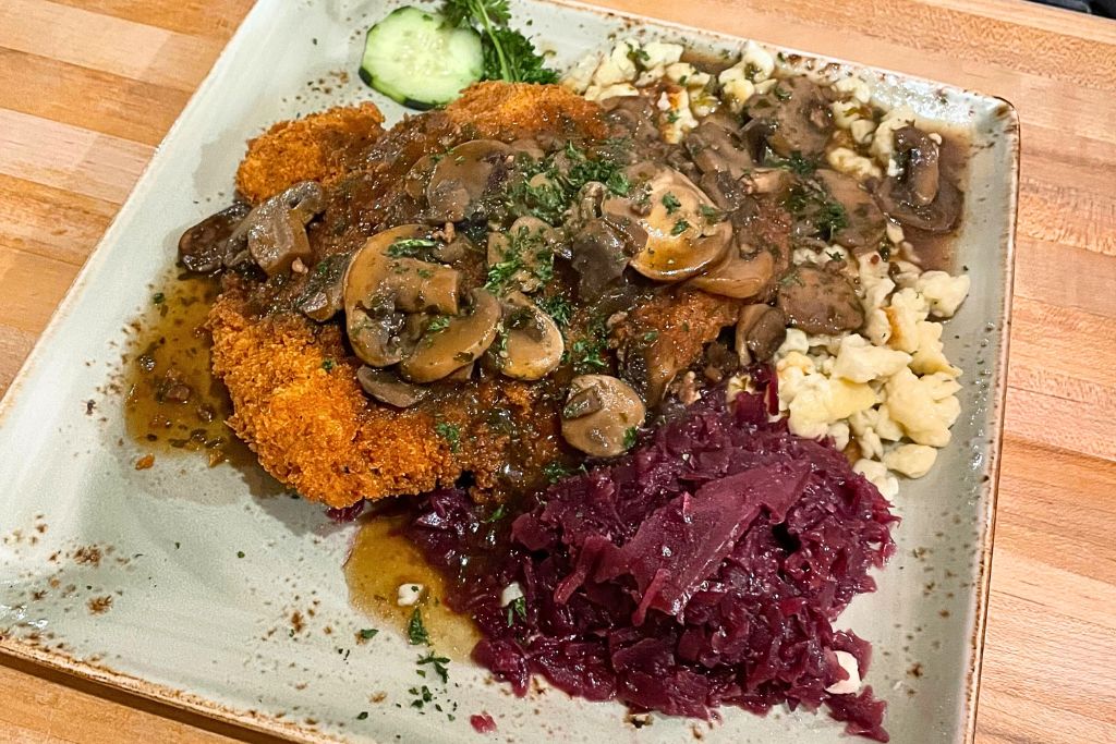 A hearty German meal featuring a breaded schnitzel topped with a mushroom gravy, served alongside spätzle, red cabbage, and a cucumber garnish. The dish is presented on a rustic plate, highlighting the rich flavors and traditional Bavarian cuisine.